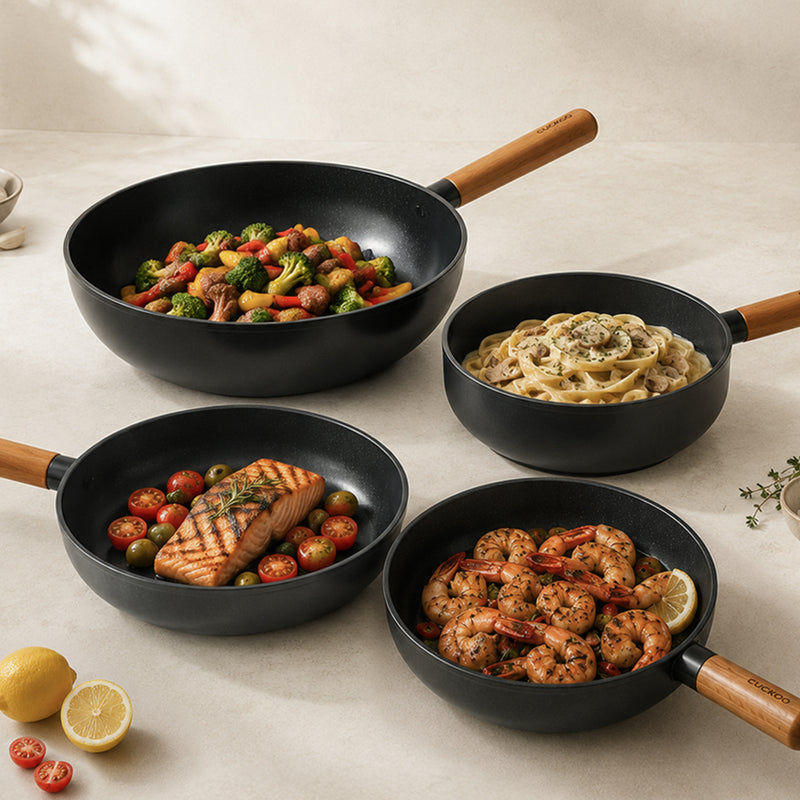 Four black nonstick frying pans with light wood handles arranged on a light beige surface, filled with cooked food including vegetables, pasta, salmon, and shrimp. Lemons and small herbs are placed nearby as styling props.