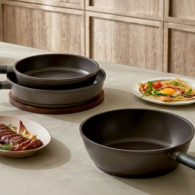Three matte black non-stick cookware pans in different sizes arranged on a light beige countertop, with a wooden cabinet background and plates of food nearby.
