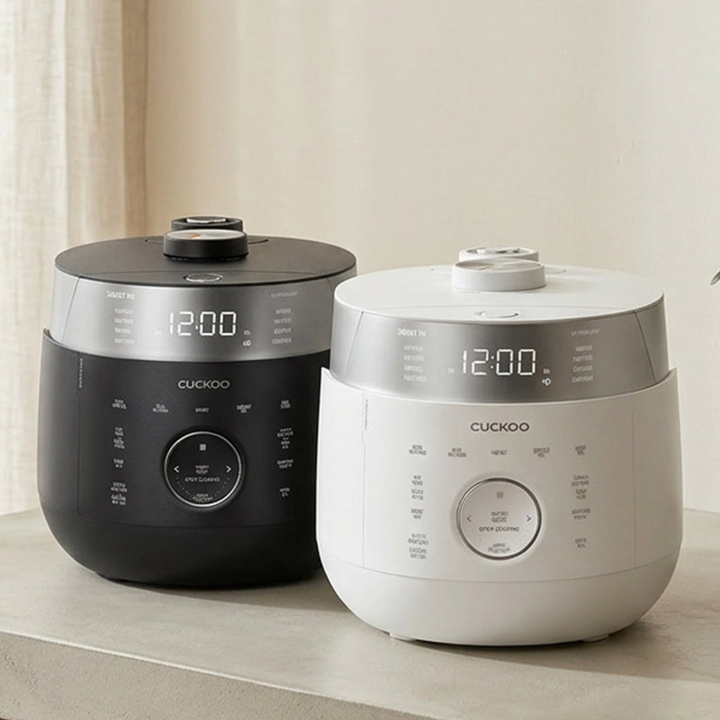 Two Cuckoo rice cookers, one in matte black and one in matte white, sitting side-by-side on a light beige countertop against a neutral cream background.