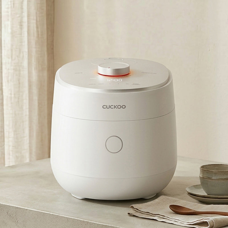 A white Cuckoo rice cooker sits on a light beige stone surface in a softly styled kitchen setting, with a curtain, ceramic cup, and wooden spoon nearby.