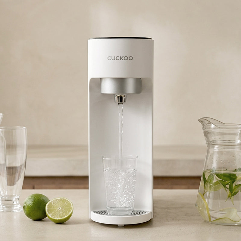 A Cuckoo water dispenser with a clear stream of water flowing into a glass, set on a countertop with limes and a glass carafe in a kitchen setting.